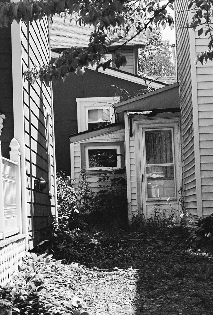 A black and white image of a view beside a house to a series of roofs, windows and doors. 