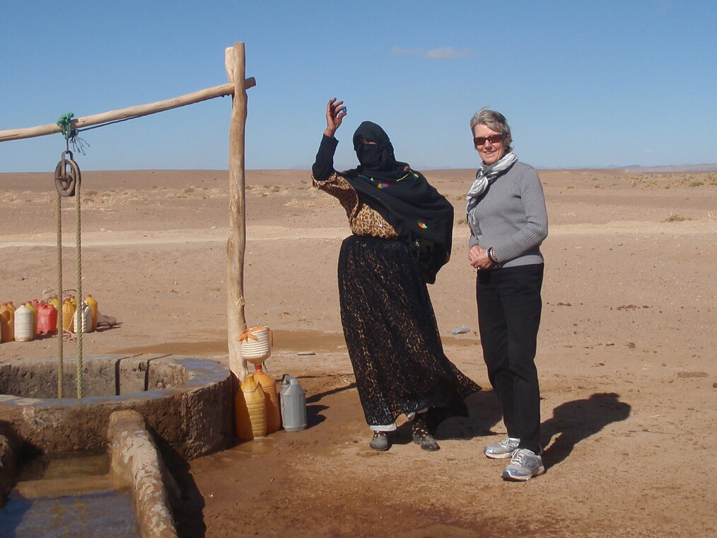 Two women are standing in the arid desert landscape of the Sahara. One is wearing a black hijab and the other is wearing a gray long sleeved shirt and black pants. The woman on the left is holding a small blue marble in her right hand, while the smiling woman on the right has her hands clasped together in front of her.