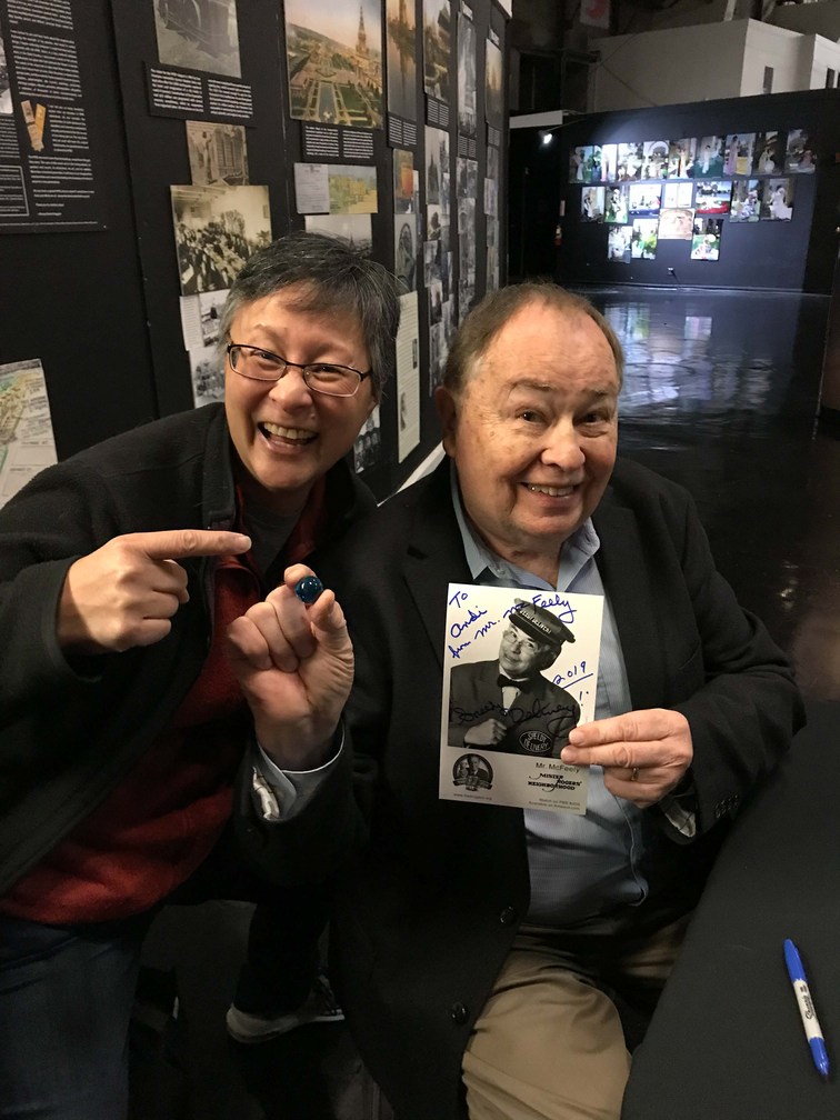 Andi Wong points at one blue marble held between the fingertips of actor David Newell. aka Mr. McFeely on Mr Roger's Neighborhood. David is seated with a signed photo of his beloved character, the friendly mailman known for his speedy and cheerful delivery of the mail.